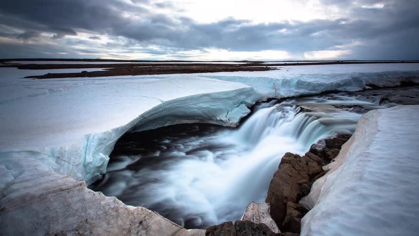 A dramatic time-lapse of a powerful waterfall flowing through a melting ice shelf under a cloudy sky in a vast arctic landscape