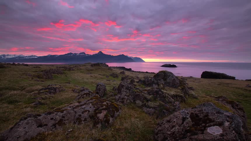 A vibrant pink and purple sunset casts a magical glow over a rocky coastline, with snow-capped mountains across the serene ocean