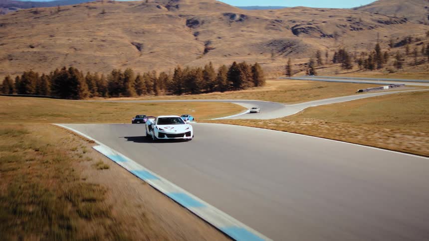 A white Corvette leads a pack of sports cars around a professional race track through a dry, hilly landscape on a sunny day
