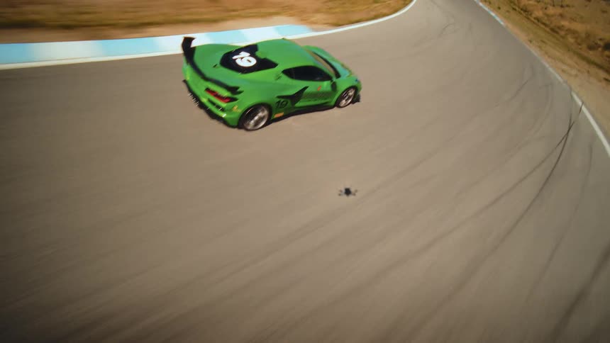 An FPV drone dynamically chases a green Chevrolet Corvette race car at high speed around a winding racetrack