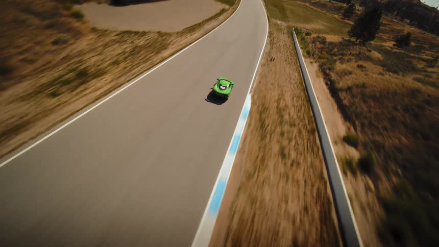 Aerial view of a green sports car racing at high speed along a straightaway on a professional track in a dry, hilly landscape
