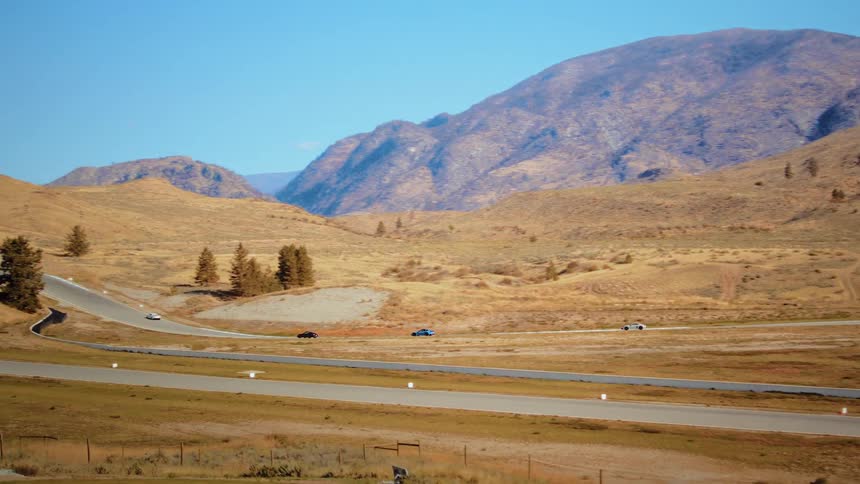 Luxury sports cars race along a winding track through a scenic, arid mountain landscape on a sunny day