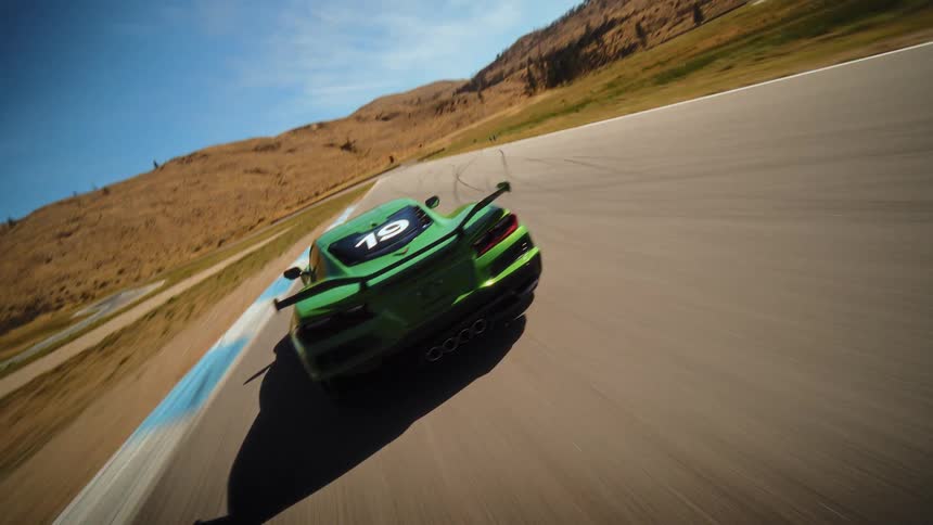 High-speed FPV drone shot follows a green Chevrolet Corvette as it speeds around a curving racetrack on a sunny day