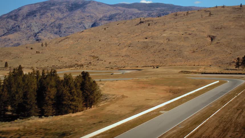 Aerial shot of a white sports car driving along a winding racetrack in a scenic, arid mountain landscape