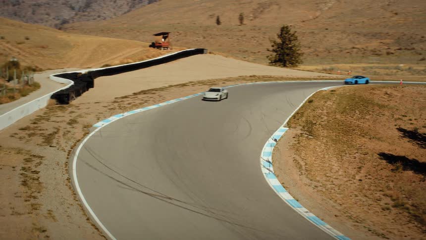 A thrilling high-angle shot captures two sports cars racing at high speed around a winding track in a scenic, arid landscape