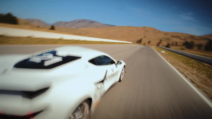 High-speed FPV drone shot follows a white Corvette number 21 as it expertly navigates a sunlit racetrack in a vast, arid landscape