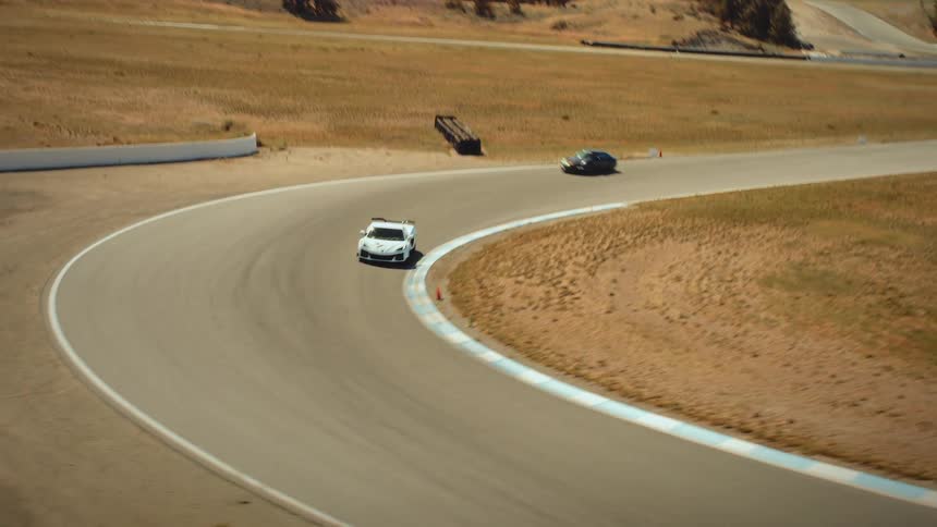 A white race car navigates a sweeping turn on a professional racetrack surrounded by dry hills and mountains