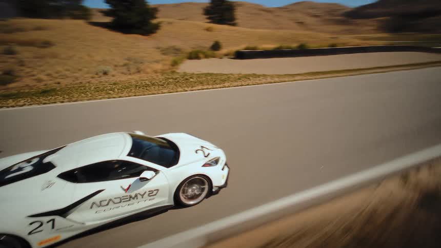A thrilling, high-speed drone chases a white Corvette as it tears around a sun-drenched race track in a hilly, arid landscape