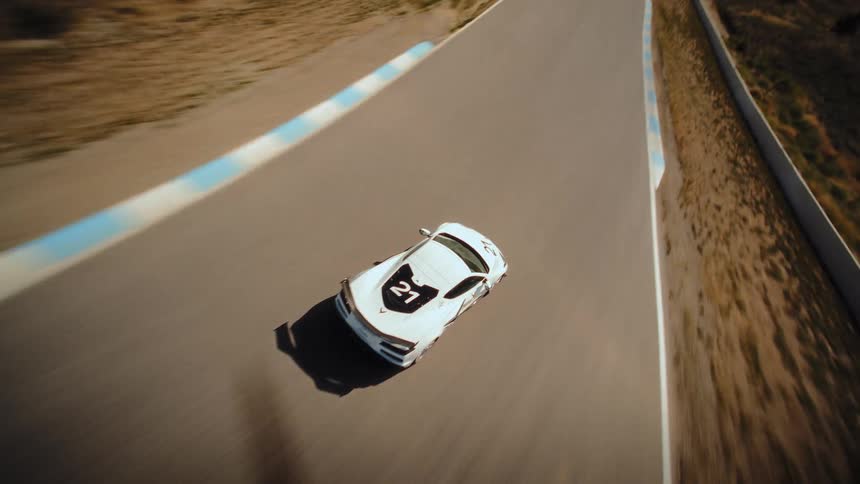 An FPV drone dynamically chases a white sports car as it speeds around a winding racetrack in a sunny, arid landscape