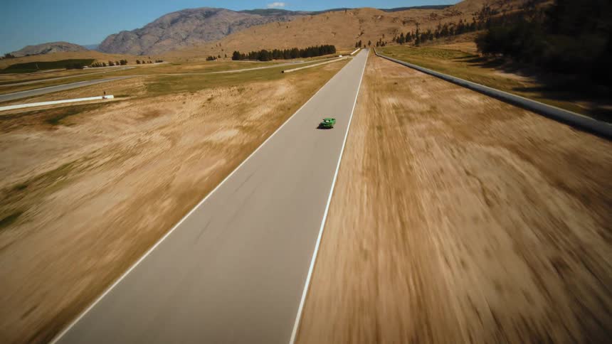 A dynamic aerial shot follows a lime green supercar as it speeds along a long straightaway on a racetrack in a vast, arid landscape
