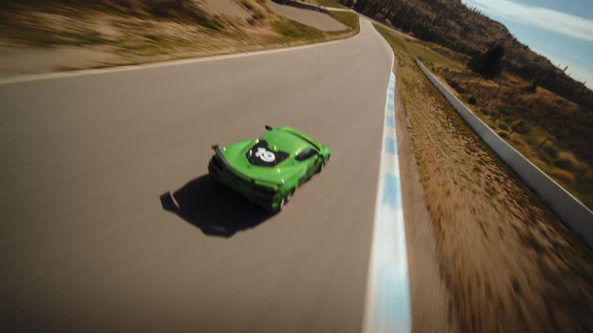 A dynamic aerial shot tracks a green sports car as it speeds around a winding racetrack through a sunlit, hilly landscape