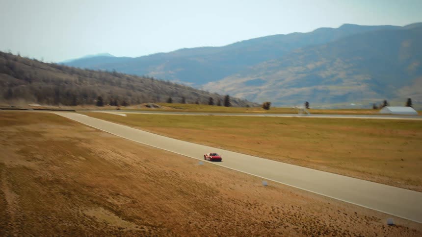 An aerial view follows a high-performance red sports car speeding down a straightaway on a racetrack through an arid, mountainous landscape