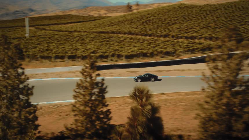Aerial tracking shot reveals a black sports car driving on a winding racetrack next to a large vineyard on a sunny day