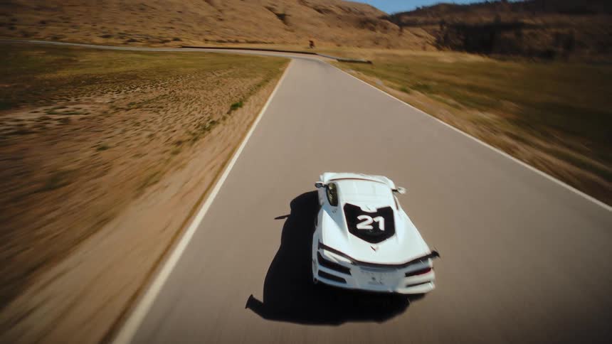 Dynamic FPV drone footage follows a sleek white sports car as it races at high speed around a winding track in a sun-drenched, hilly landscape