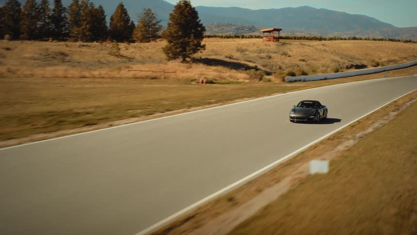 Aerial tracking shot of a sleek grey convertible sports car speeding along a racetrack through a scenic, sun-drenched landscape
