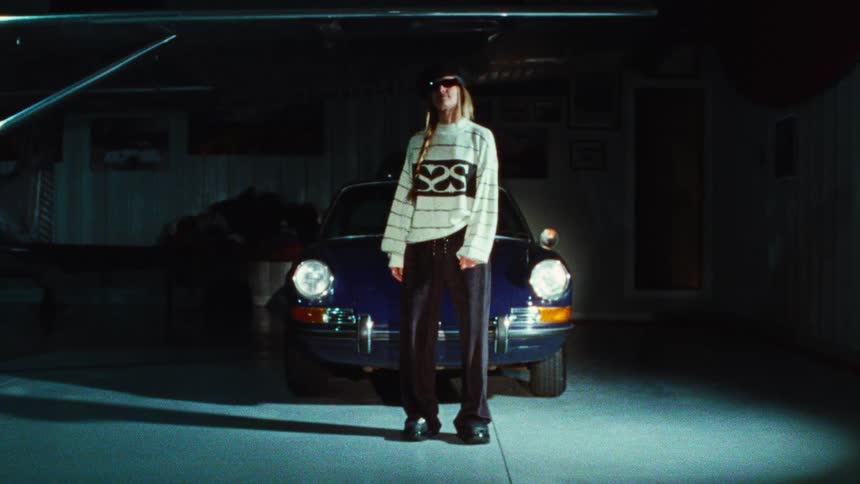 A stylish young woman in a beret and sunglasses poses in front of a classic blue sports car in a dimly lit airplane hangar