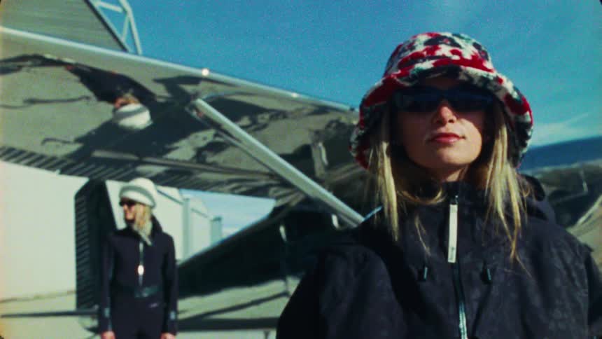 Vintage 8mm footage of two stylish women in sunglasses posing in front of a small prop plane