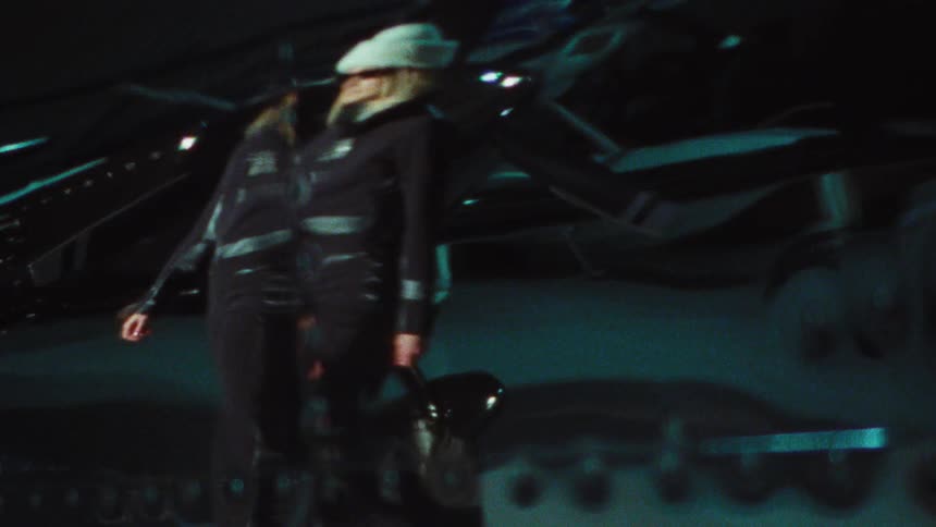 A pilot in a jumpsuit and hat walks past a dark aircraft in a hangar with a distorted, dreamlike effect
