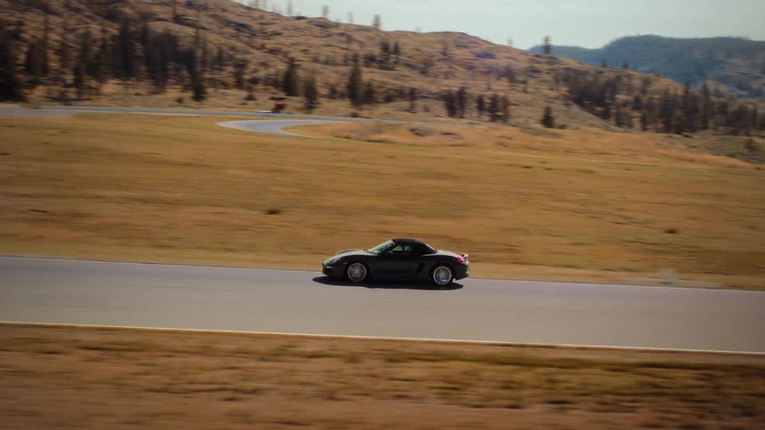 Side view tracking shot of a black Porsche convertible speeding along a racetrack in a scenic, arid mountain environment