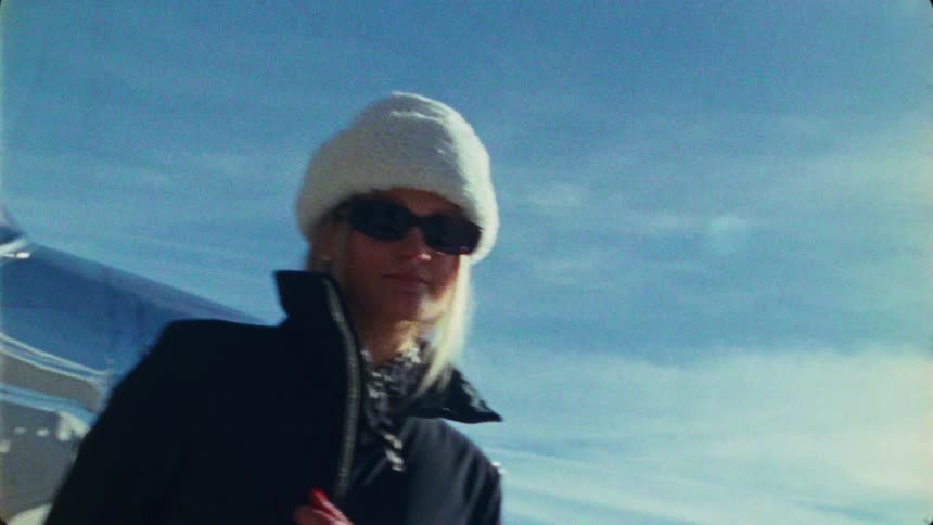 Vintage 16mm film of a stylish blonde woman in a winter hat posing in front of a propeller airplane