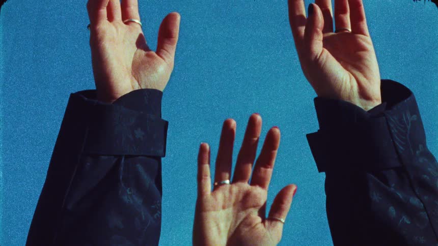 Three hands with rings reach up towards a vibrant blue sky in a grainy, retro-style film clip