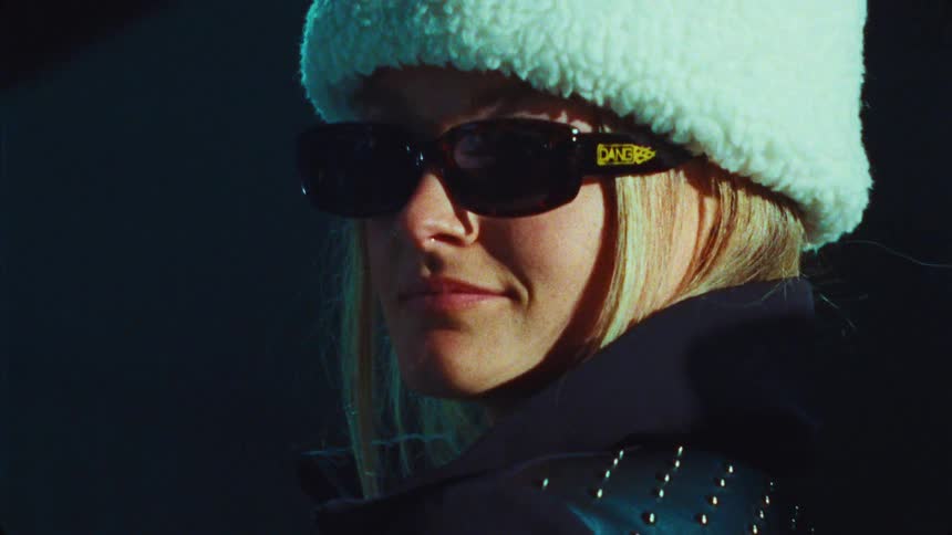 A close-up portrait of a stylish blonde woman in a fuzzy white hat and sunglasses, smiling with a confident look on grainy vintage film
