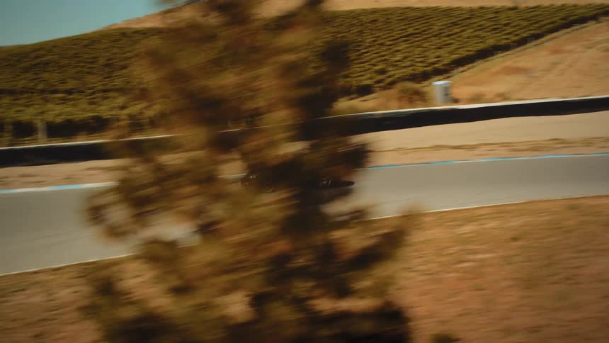 A high-angle tracking shot follows a sleek black sports car as it speeds around a winding racetrack set in a dry, hilly landscape