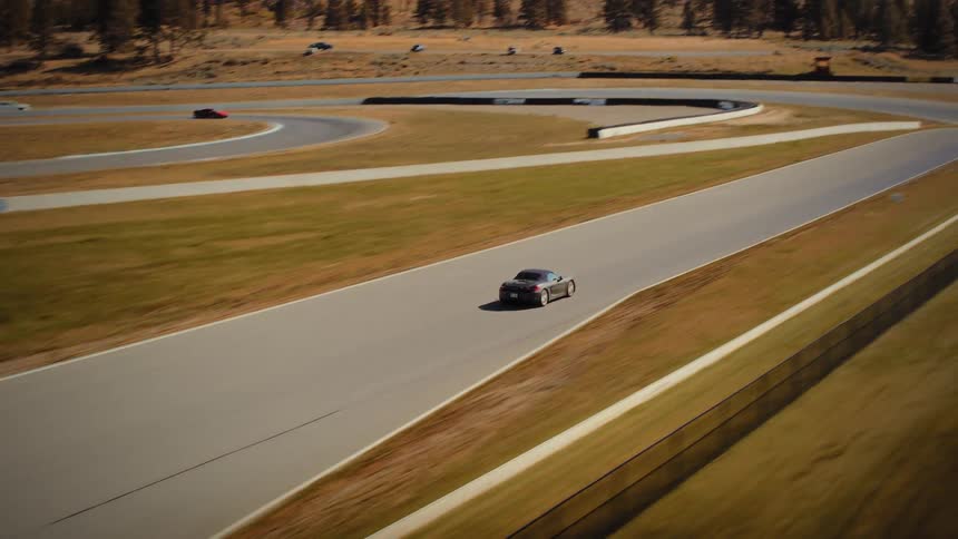 High-angle aerial tracking shot follows a grey convertible sports car as it speeds down the main straight of a rural race track on a sunny day