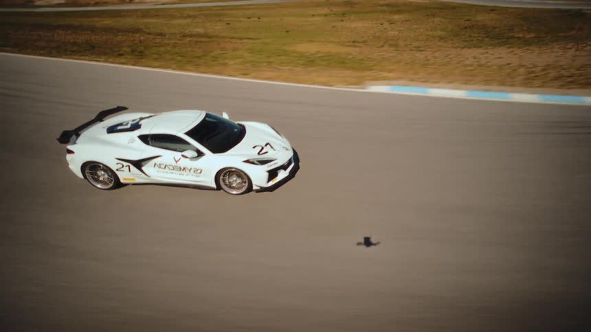 A fast-moving FPV drone captures dynamic aerial footage as it chases a white Corvette race car around a desert racetrack