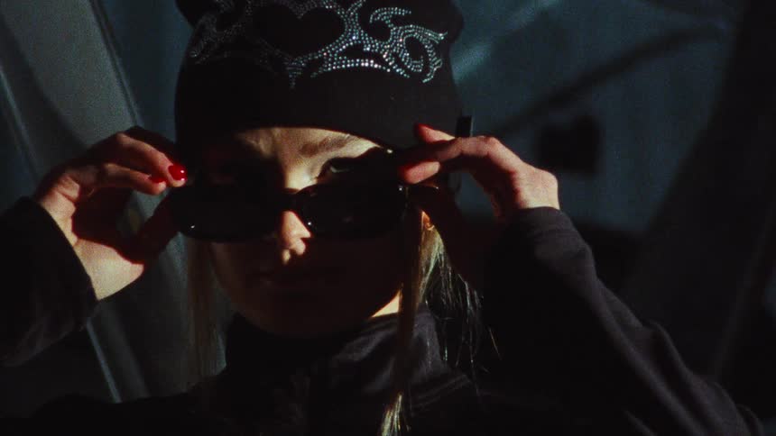 A stylish woman in a black beanie slowly puts on her sunglasses, looking off-camera with a cool expression in a dimly lit airplane hangar