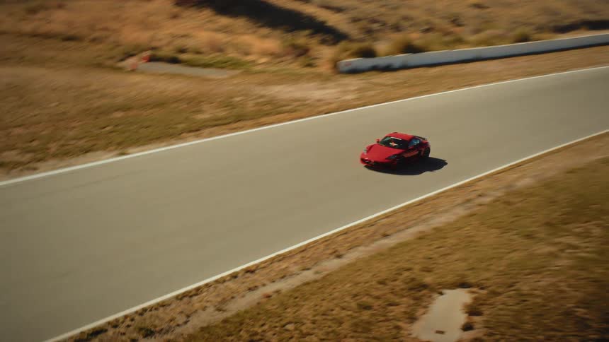 An aerial view shows a red sports car speeding along a race track in a dry, rural landscape