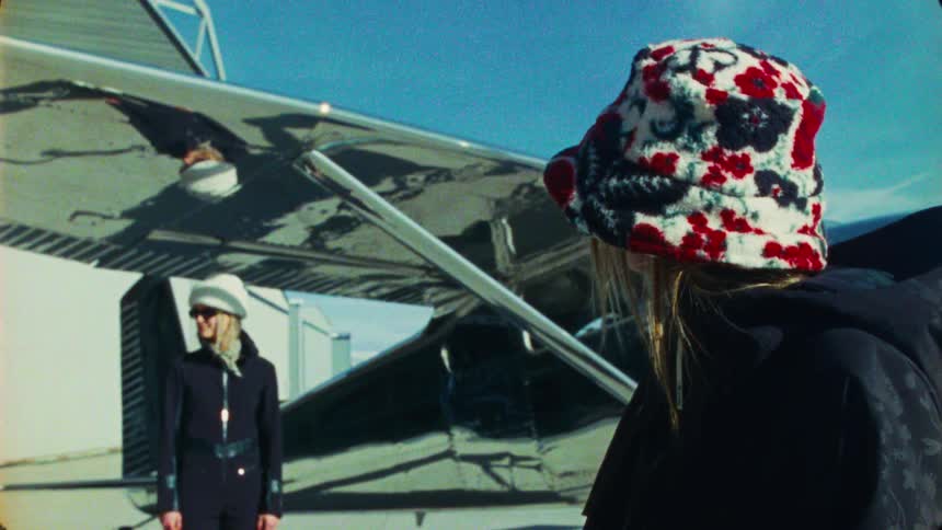 A stylish woman in a colorful bucket hat and sunglasses poses in front of a reflective airplane on a sunny day