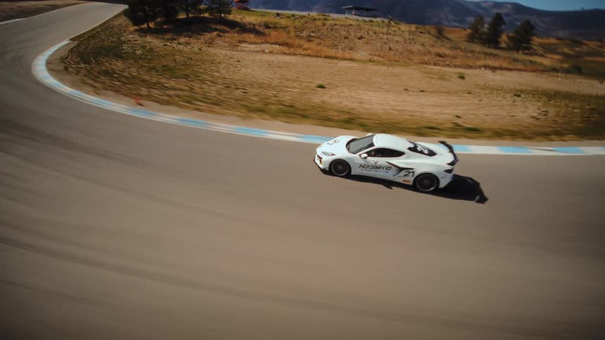 An FPV drone captures thrilling high-speed footage as it chases a white Corvette race car around a winding professional racetrack