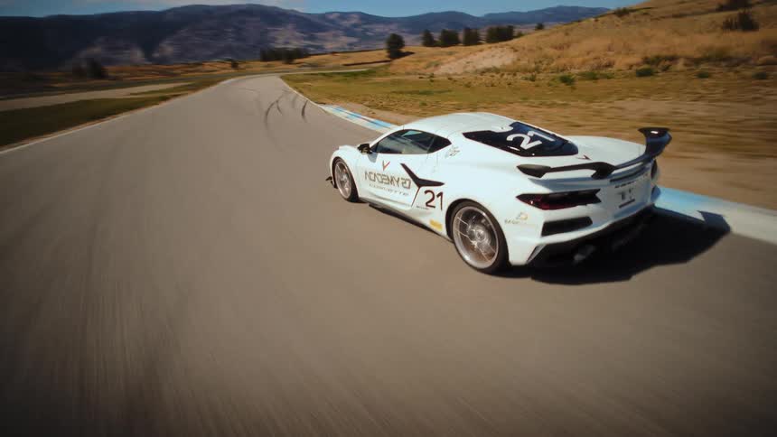 High-speed FPV drone footage chases a white Chevrolet Corvette race car as it speeds around a curvy track