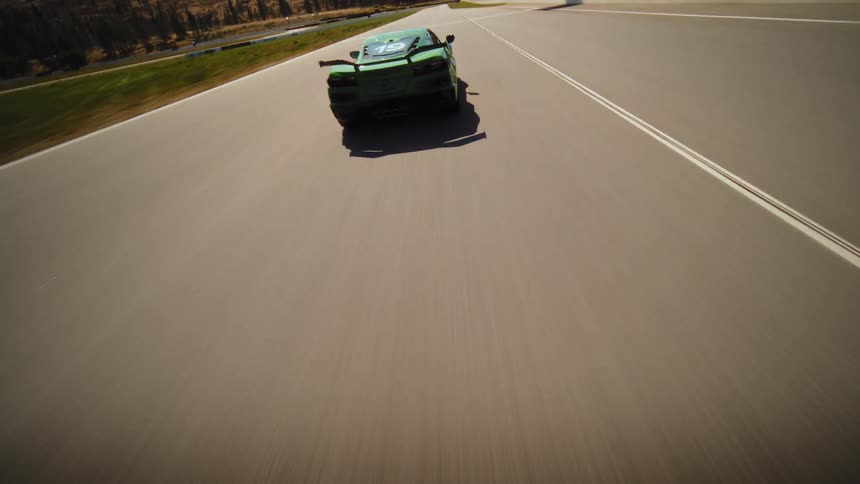 A dynamic FPV drone chases a green sports car as it speeds around a sunny professional race track in the mountains