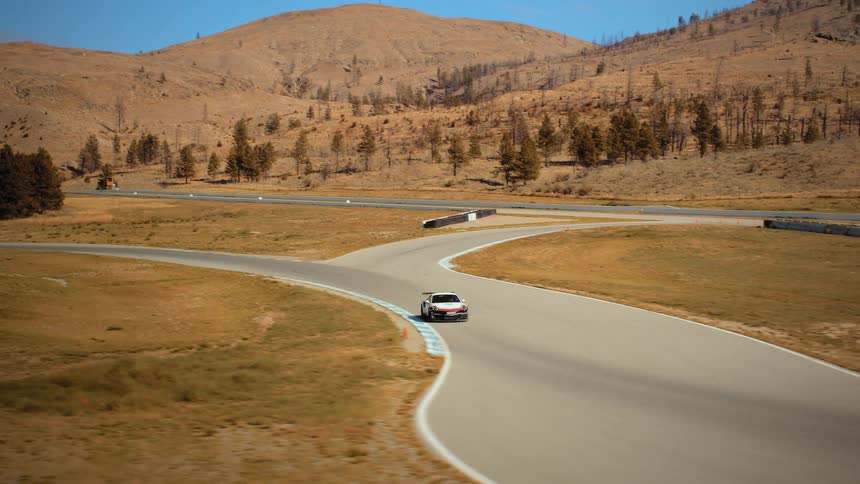 A white race car speeds around a winding track through a dry, mountainous landscape on a sunny day