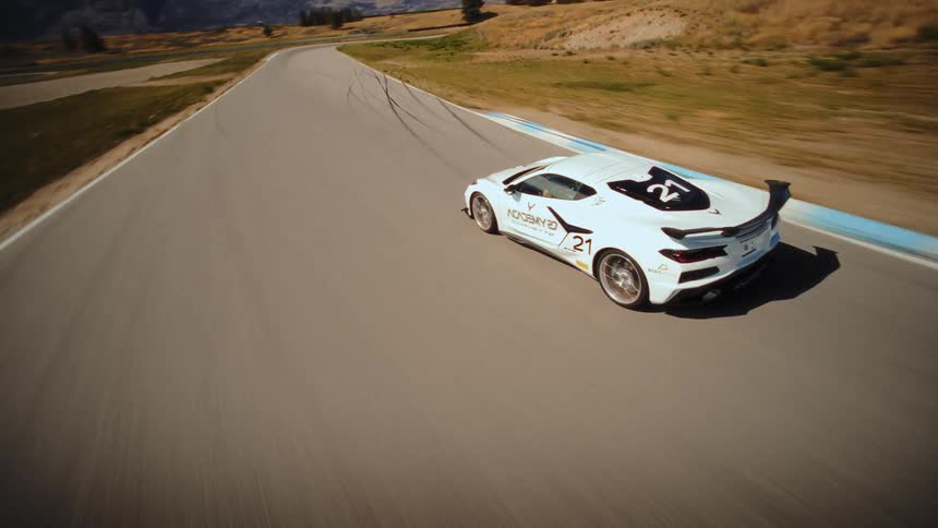 A dynamic FPV drone chases a white Corvette race car at high speed around a winding track in a sunny, arid landscape