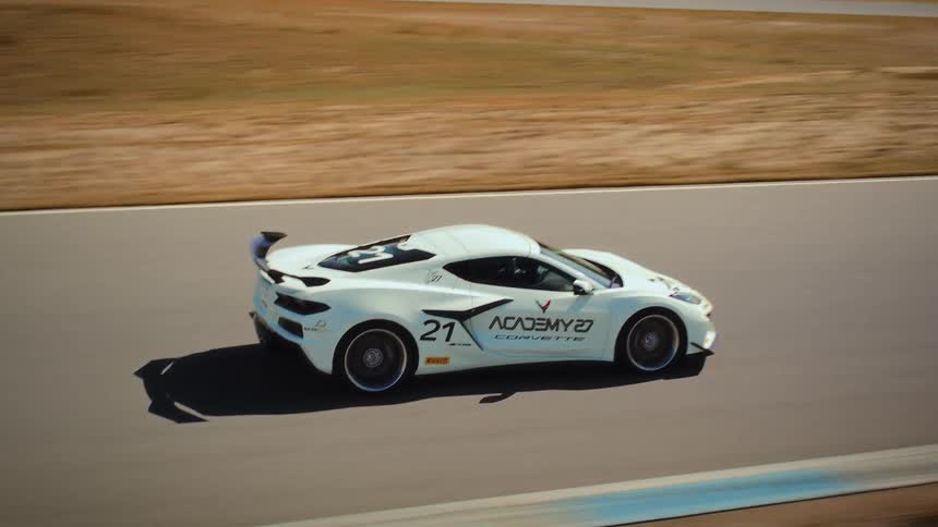 Dynamic aerial view of a white Chevrolet Corvette racing around a paved track surrounded by dry hills