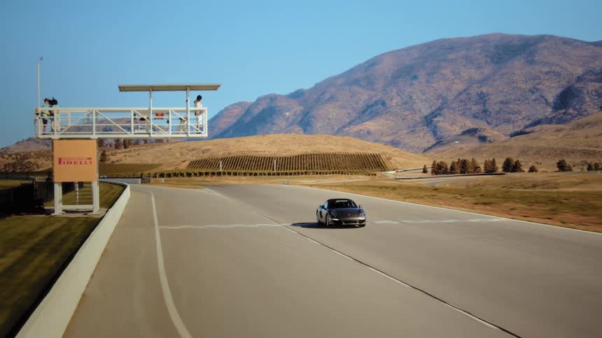 A dark luxury convertible sports car speeds around a scenic race track surrounded by arid, sun-kissed mountains and hills