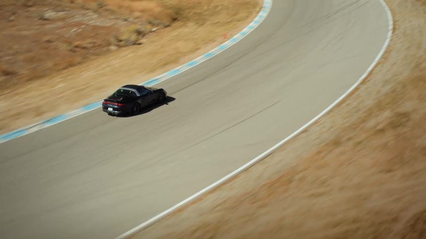 Cinematic aerial tracking shot of a black luxury sports car driving along a racetrack in a dry, rural landscape