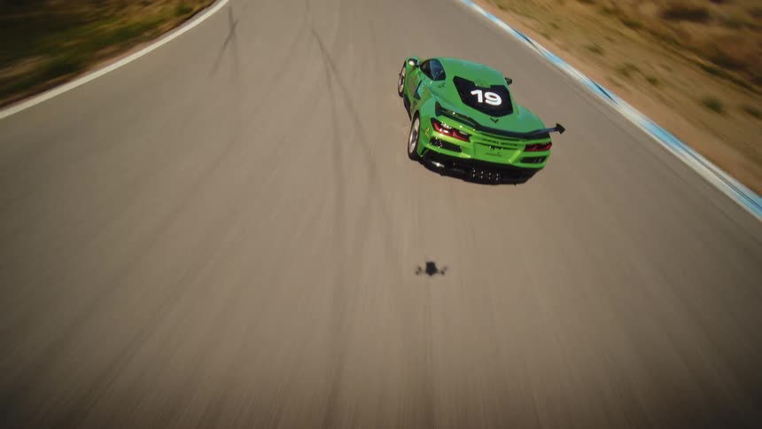 A thrilling FPV drone chase follows a high-performance green race car as it speeds around the corners of a track in a dry, hilly landscape