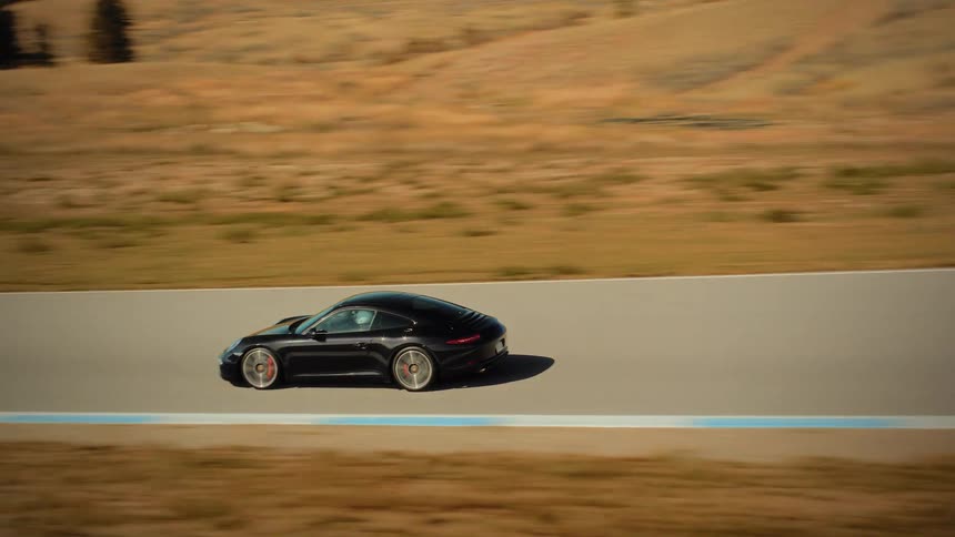 A sleek black sports car gracefully navigates a turn on a sunny racetrack set in a dry, rolling hills landscape