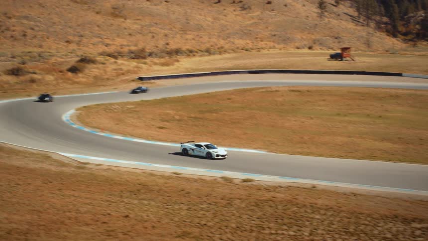An aerial tracking shot follows a white sports car as it races along a winding track through a dry, hilly landscape