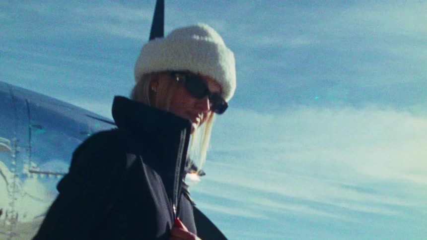 A stylish blonde woman in a fuzzy hat and sunglasses poses in front of a vintage silver airplane on a sunny day