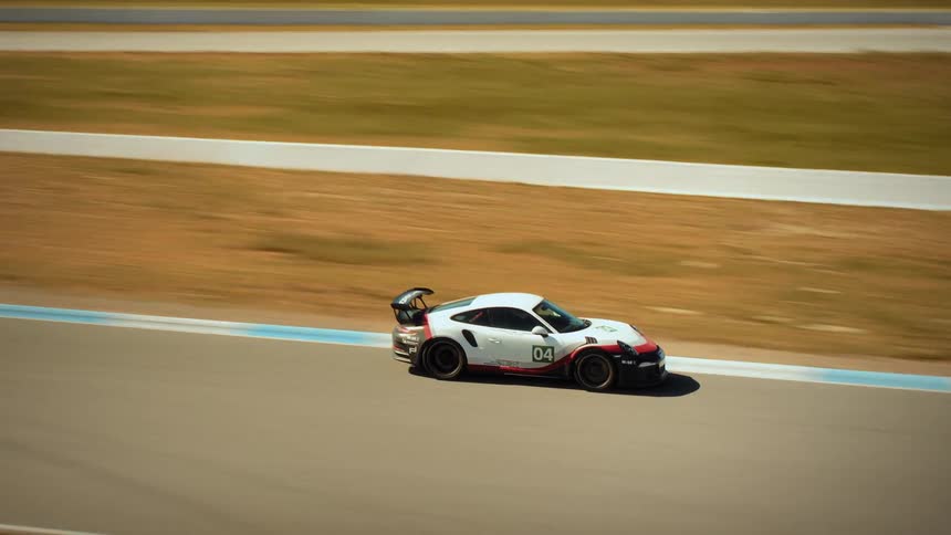 Aerial tracking shot follows a white Porsche GT3 race car as it speeds along a professional racetrack in a dry, hilly landscape