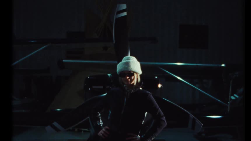 Vintage-style footage of a fashionable female aviator posing confidently in front of her aircraft in a dark hangar at night