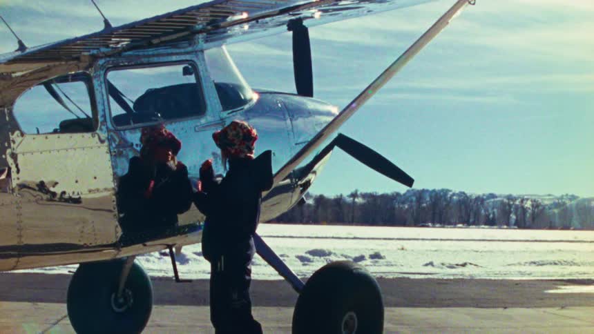 Vintage 16mm film shows two people in winter gear standing next to a chrome propeller plane on a snowy airfield