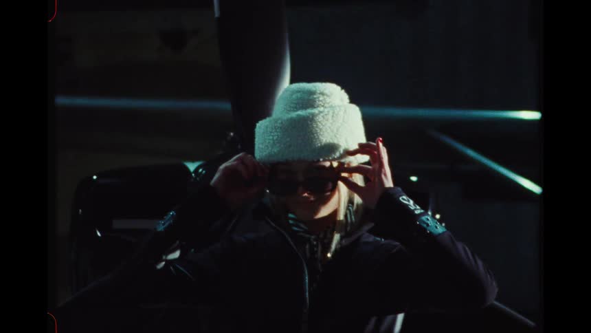 A stylish woman in a white hat and sunglasses poses in front of a helicopter in a hangar, shot on vintage film
