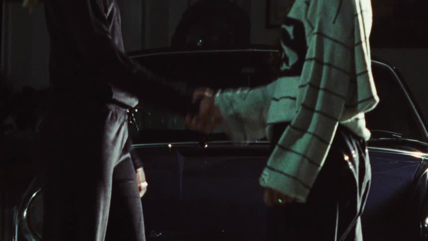 Two people finalize a deal with a firm handshake in front of a vintage car in a dark garage