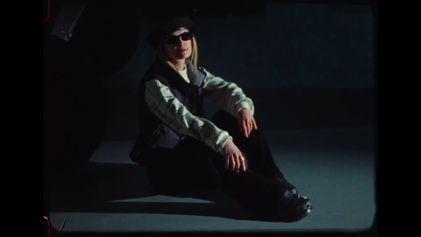 Stylish woman in a beret and sunglasses sits in a spotlight during a fashion photoshoot on vintage film